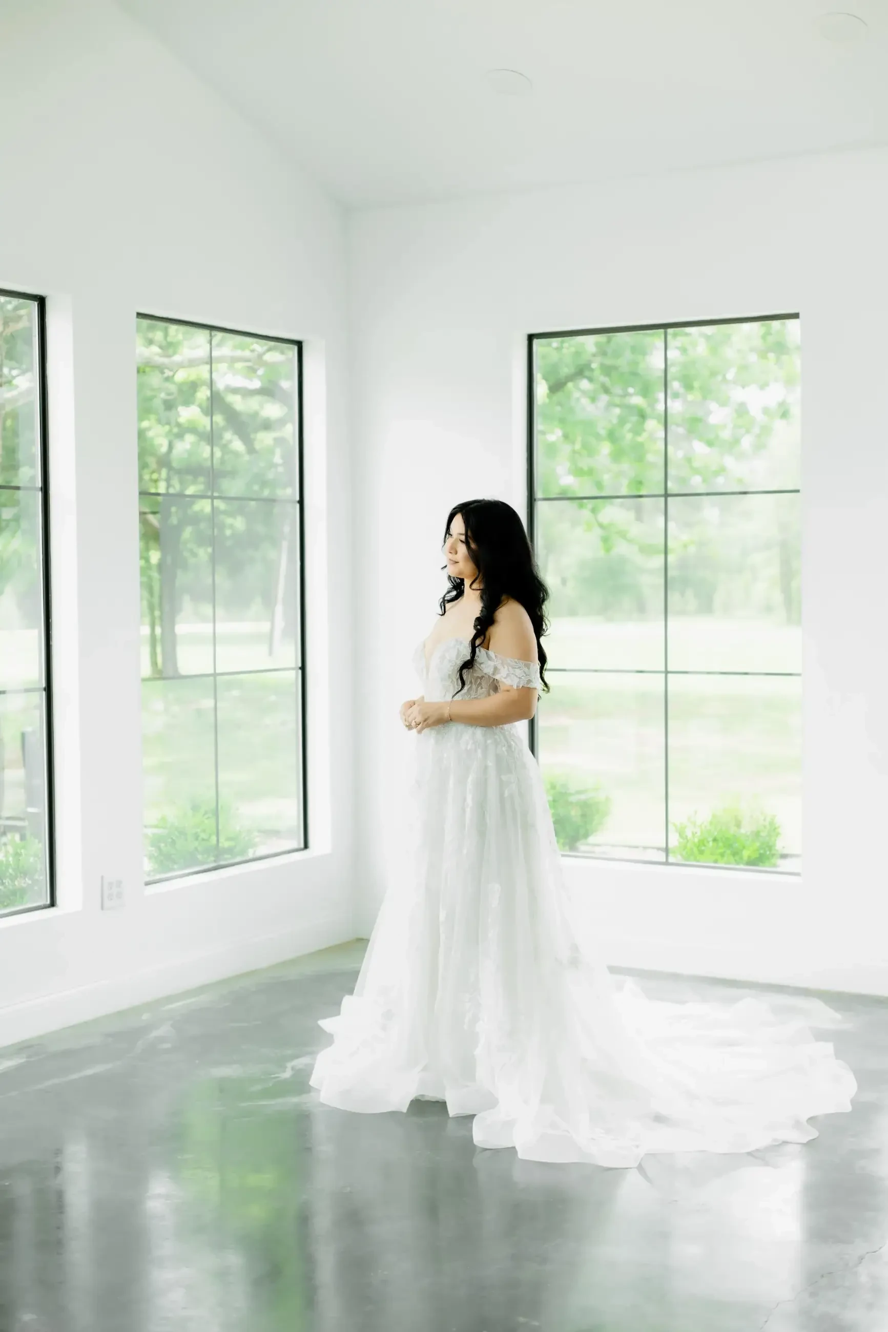 A bride in an off-shoulder, lacy white gown stands in a bright room with large windows. She looks out thoughtfully. The space feels serene and elegant.